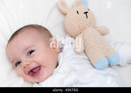 BABY WITH DUMMY. Little baby boy resting on the bed happy and smiling with a bunny dummy made of crochet fabric aside. Top view. Stock Photo