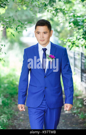 Male model, the groom portrait in a blue suit with a boutonniere among the green foliage on a wedding day. Stock Photo