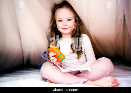 Little girl reading fairy tales book under the covers at the evening with lantern. Stock Photo