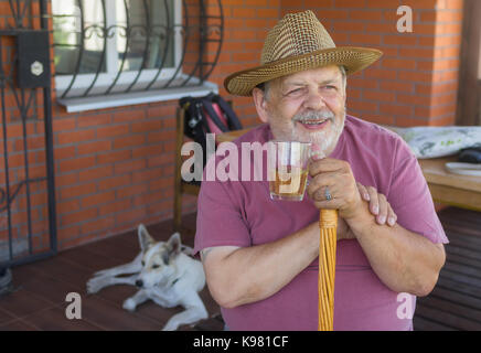 Portrait of bearded smiling senior man holding glass of tea while sitting on veranda near his house Stock Photo