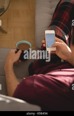 Man using mobile phone while having cup of coffee in living room Stock Photo