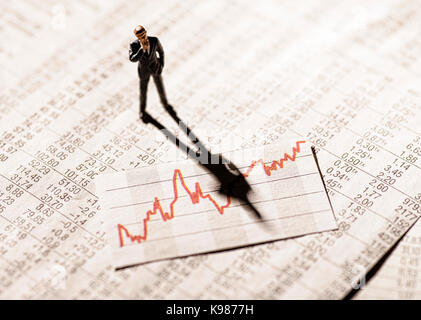 Model figure stands on rate tables and looks skeptically on a graph with stock prices. Stock Photo