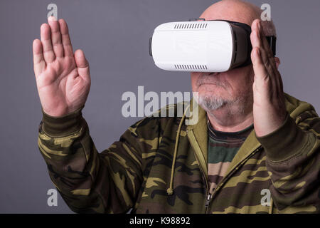 Senior man in wearable technology VR glasses. Confident old man wearing camouflage clothing in virtual reality headset with Interface selective focus  Stock Photo