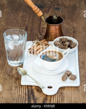 Cup of black coffee, copper pot, water with ice in glass, cinnamon sticks and cane sugar cubes on white ceramic serving board over rustic wooden backdrop. Stock Photo