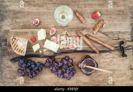 Glass of white wine, cheese board, grapes, figs, strawberries, honey and bread sticks on rustic wooden background Stock Photo