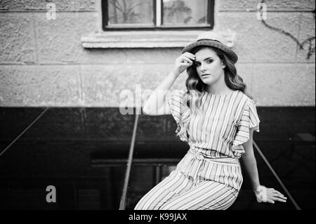 Portrait of a fabulous young woman wearing striped overall and hat sitting on black shiny surface next to the building. Black and white photo. Stock Photo
