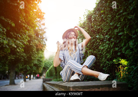 Portrait of a stunning young woman in striped overall sitting in the park and listening to the music with her earphones on. Stock Photo