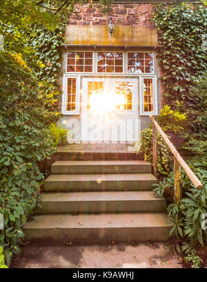 Sunbursts gleam through glass doors surrounded by lush green foliage and ivy on a weathered window and old stone wall Stock Photo