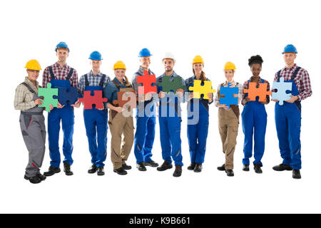 Portrait of confident construction workers holding jigsaw pieces while standing in row against white background Stock Photo