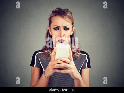 Closeup portrait funny shocked scared woman looking at phone seeing bad news photos message with disgusting emotion on face isolated on gray backgroun Stock Photo