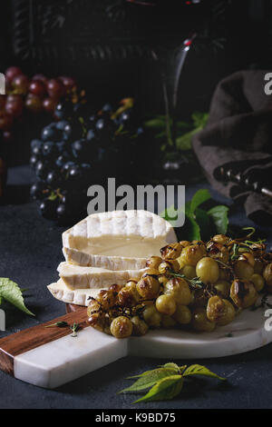 Serving board with sliced camembert cheese and baked bunch of green grapes served with glass of red wine, corkscrew, green leaves over black table. Da Stock Photo