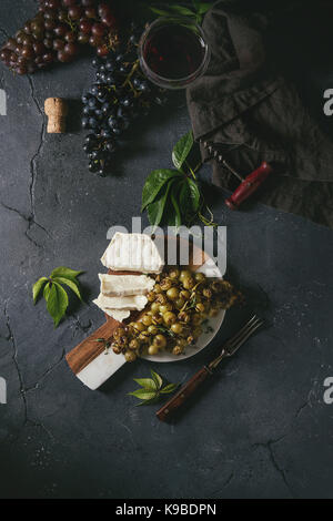 Serving board with sliced camembert cheese and baked bunch of green grapes served with bread, glass of red wine, corkscrew, green leaves, fork over bl Stock Photo