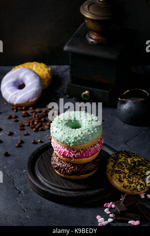 Variety of colorful glazed donuts with chopped chocolate, pink sugar, coffee beans vintage coffee grinder on black texture background. Dark rustic sti Stock Photo