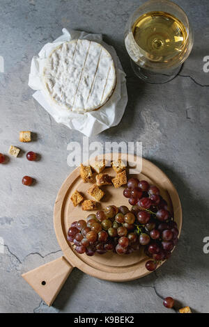 Bunch of red grapes, camembert cheese, croutons and glass of white wine served on wooden serving board over gray kitchen table. Rustic style. Top view Stock Photo