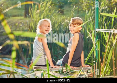 Summertime in the countryside. Two children (sibling or best friends) sitting on the pier of the lake. Stock Photo