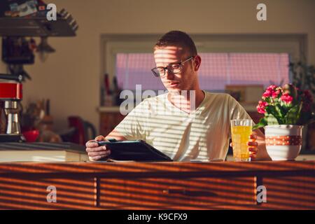 Morning time at home. Young man reading on the digital tablet in the kitchen during breakfast. Stock Photo