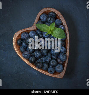 Fresh blueberries with a leaf of mint in the heart-shaped bowl on a dark blue background Stock Photo