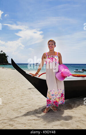 A beautiful Filipino woman poses by an outrigger in her flowery dress and pink wide-brim hat on White Beach near Puerto Galera, Mindoro, Philippines. Stock Photo