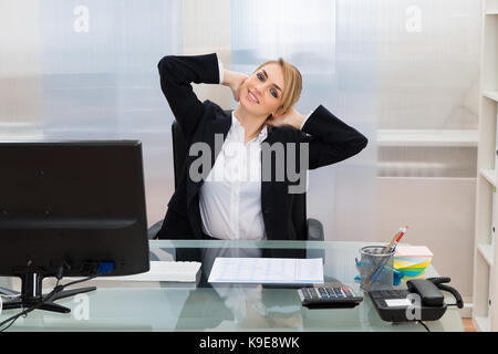 Young Happy Businesswoman With Hands Behind Head Sitting At Desk Stock Photo