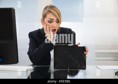 Shocked Businesswoman Holding Broken Screen Digital Tablet In Office Stock Photo