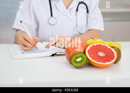 Close-up Of Female Dietician Writing Prescription With Fruits On Desk Stock Photo