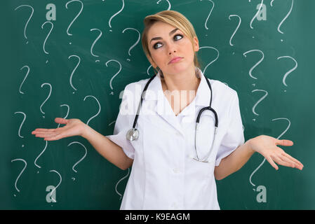Female Doctor Shrugging In Front Of Blackboard With Question Mark Signs Stock Photo