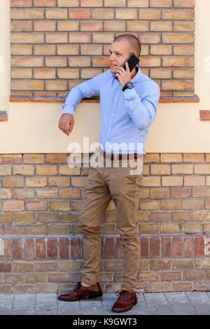 Young handsome businessman standing in front of the building and talking on a mobile phone. He arranges a meeting with his new business partners via a Stock Photo