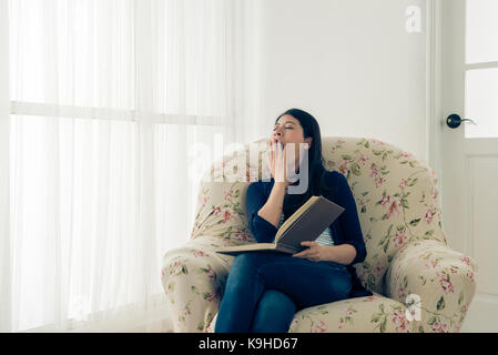 vintage retro film color photo of pretty beauty female model sitting on comfortable sofa reading diary book feeling tired yawning in living room at ho Stock Photo