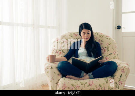 vintage retro film color photo of attractive happy woman reading diary book and holding hot coffee mug sitting in front of window comfortable sofa at  Stock Photo