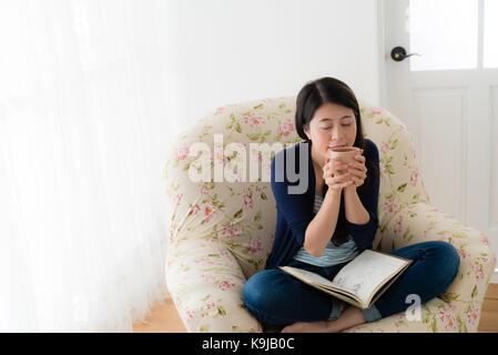 lovely beautiful female model holding hot coffee cup enjoying espresso sitting on living sofa reading at home with high angle view photo. Stock Photo