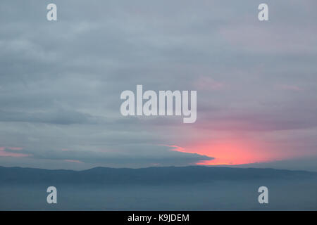 Red sunset filtering through an hole in the overcast sky, with mist filling a valley below Stock Photo