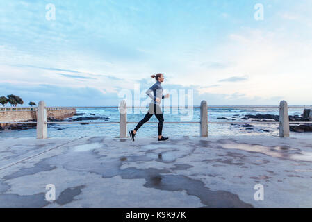 Woman athlete running outdoors in the morning. Young female runner running on road along the sea. Stock Photo