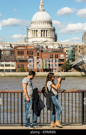 Young Couple Leaning Against a Fence By The River Thames in Bankside Southwark London Wearing Grey Tops and Blue Denim Jeans Taking Photographs With a Stock Photo