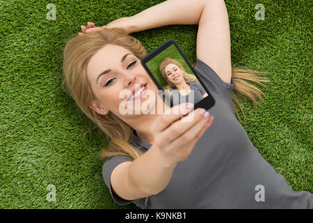 Happy Woman Lying On Grass Taking Selfie With Mobile Phone Stock Photo