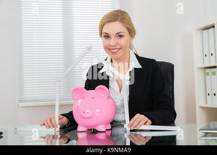 Happy Businesswoman Holding Ruler Folded In House Shape With Piggy Bank At Desk Stock Photo