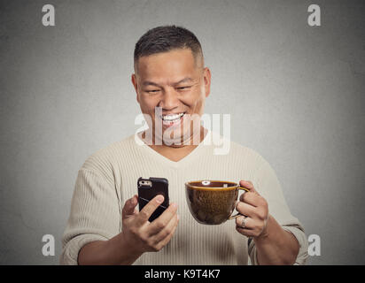 Closeup portrait happy, smiling middle aged man reading good news on smart phone, holding mobile, drinking cup coffee isolated grey background. Human  Stock Photo