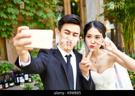happy young asian bride and groom making a face while taking a selfie using cellphone. Stock Photo