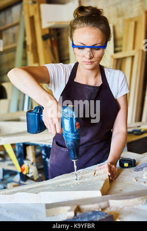 Portrait of young woman working in carpenters shop drilling holes in piece of wood while making furniture Stock Photo