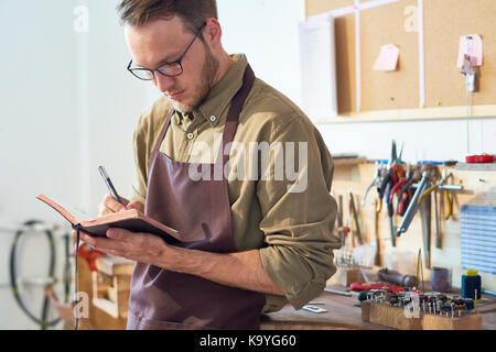 Portrait of young craftsman drawing sketches holding open book while standing in workshop against table with tools Stock Photo