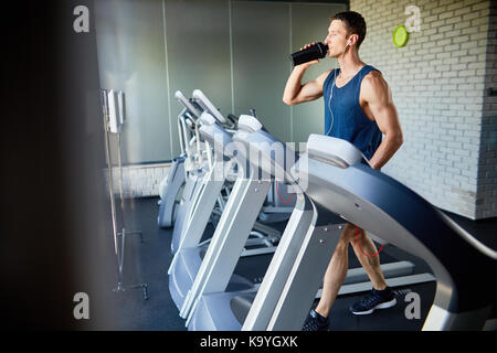 Young sportsman in headphones drinking water from bottle while running on treadmill in modern gym, profile view Stock Photo