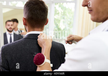 Confident young tailor standing behind his male customer and measuring width of shoulder for custom-made garment, interior of modern sewing atelier on Stock Photo