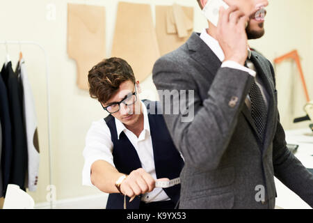 Creative young tailor taking back measurements of busy client in order to make classical suit, interior of modern sewing atelier on background Stock Photo