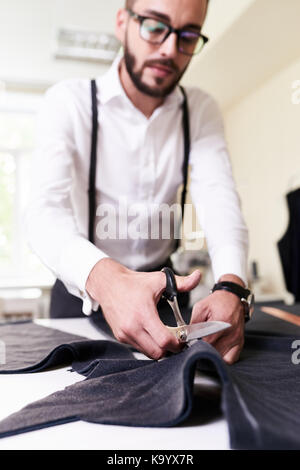Concentrated young designer in eyeglasses cutting fabric with scissors in order to make male jacket, interior of modern studio on background Stock Photo