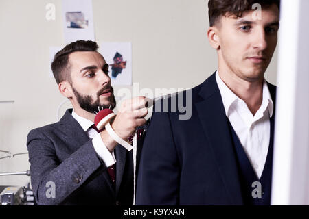 Handsome young tailor with stylish haircut taking measurements of customer in order to make classical suit, interior of modern studio on background Stock Photo