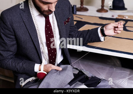 Bearded young tailor wrapped up in sewing male jacket with needle and thread while sitting at working table, interior of atelier on background Stock Photo