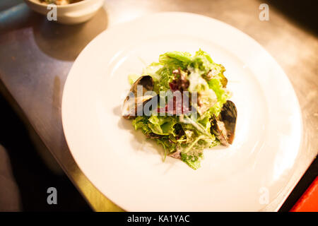 The close-up side view of the seafood salad consisted of the lettuce, sausage and mussels placed on the white plate. Stock Photo