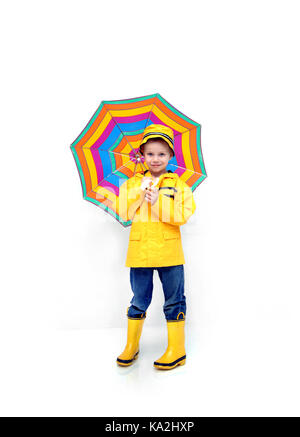 Little boy stands prepared for rain.  He is wearing a yellow raincoat and hat.  He is carrying a striped umbrella and has his jeans tucked into yellow Stock Photo