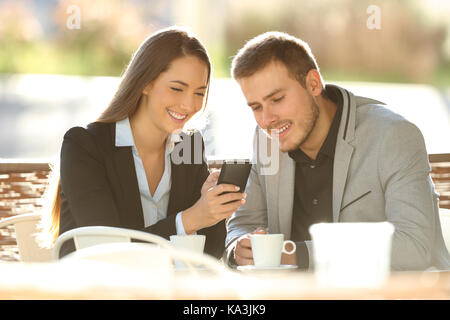 Two happy executives using a smart phone sitting in a restaurant terrace with a warm light in the background Stock Photo