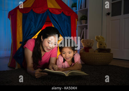 pleasantly beautiful housewife and pretty sweet children lying on toy tent reading story book together in bed room at night. Stock Photo