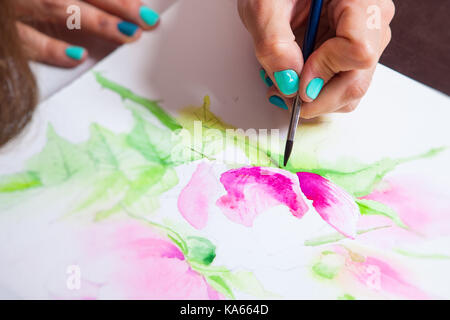 Close-up of the artist draws watercolor in the album for drawing with a thin wooden brush, on the table a pallet for drawing and a mug with tools Stock Photo